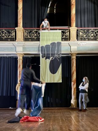 Eine Person hängt ein großes Banner mit dem Symbol einer schwarzen Faust von einem Balkon, während andere, alle mit Masken, auf der Bühne darunter stehen und arbeiten.
