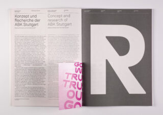 Eine aufgeschlagene Zeitschrift zeigt auf der linken Seite einen deutsch-englischen Artikel und auf der rechten Seite einen großen, fettgedruckten Buchstaben „R“ auf grauem Grund, darunter ist teilweise eine rosa Seite zu sehen.