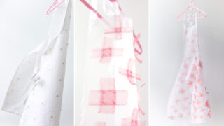 Drei durchsichtige weiße Kleidungsstücke mit unterschiedlichen rosa und orangefarbenen Mustern – Kreise, Kreuze und Gitter – werden auf Kleiderbügeln vor einem weißen Hintergrund präsentiert.