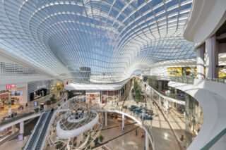 Geräumiges, modernes Einkaufszentrum mit Glasdecke, großem zentralen Atrium, mehreren Geschäften, Gehwegen und einer geometrischen Kunstinstallation.