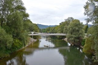 Eine moderne weiße Fußgängerbrücke überquert einen ruhigen Fluss, der von grünen Bäumen umgeben ist. Auf der Brücke gehen Menschen spazieren, im Hintergrund sind Hügel zu sehen.
