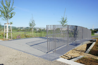 Eine kleine moderne Fußgängerbrücke mit Metallgeländer überquert eine flache, begrünte Fläche in einem Park mit jungen Bäumen.