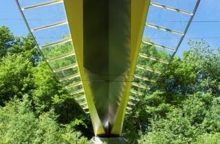 Unterseite einer modernen gelben Fußgängerbrücke mit Glasgeländer, umgeben von grünen Bäumen und Laub.