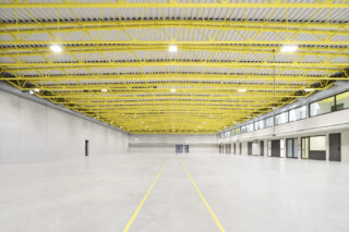 Großes leeres Lagerhaus mit Betonböden, grauen Wänden, gelben Deckenbalken und Fenstern und Türen auf der rechten Seite; auf dem Boden sind gelbe Linien markiert.