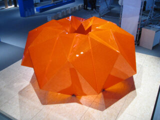 Eine große, geometrische orangefarbene Skulptur mit mehreren eckigen Facetten, die auf einer Sperrholzplattform in einem Ausstellungsraum im Innenbereich ausgestellt ist.