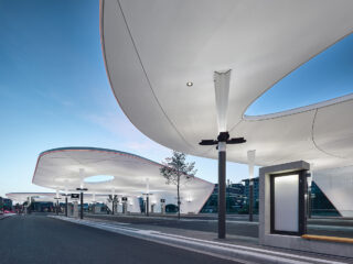 Ein moderner Busbahnhof mit großen, geschwungenen weißen Vordächern, schlanken Stützsäulen und minimalistischen Bushaltestellen in der Abenddämmerung.