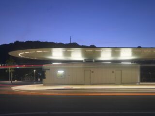 Ein moderner, runder Busbahnhof mit beleuchtetem Überhang in der Dämmerung; im Vordergrund sind Lichtspuren vorbeifahrender Fahrzeuge sichtbar.