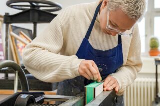 Eine Person, die eine Brille und eine blaue Schürze trägt, richtet ein buntes Buch auf einer Werkbank in einer Werkstatt sorgfältig aus.