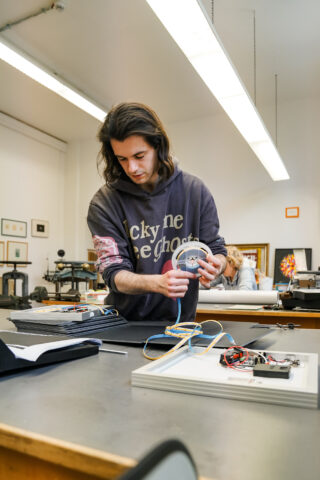 Eine Person steht an einem Tisch und arbeitet in einem gut beleuchteten Klassenzimmer oder einer Werkstatt mit elektronischen Bauteilen und Kabeln.
