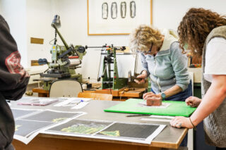 Drei Personen arbeiten in einem Atelier; eine von ihnen schlägt mit einem Hammer auf ein ziegelsteinähnliches Objekt, während auf einem Tisch im Vordergrund gedruckte Bilder und Werkzeuge ausgebreitet sind.
