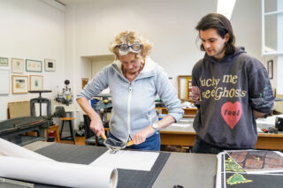 Eine ältere Frau benutzt einen Papierschneider, während ein jüngerer Mann in einem Kunstatelier mit verschiedenen Werkzeugen und Drucken an den Wänden zusieht.