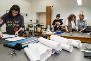 Vier Personen arbeiten an verschiedenen Aufgaben in einer Werkstatt; einer schneidet Papier zu, während die anderen mit Werkzeugen und Materialien hantieren, wobei auf einem Tisch im Vordergrund aufgerollte Papiere liegen.