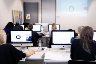 Eine Gruppe von Menschen sitzt in einem Klassenzimmer an Schreibtischen und benutzt iMac-Computer. Auf ihren Bildschirmen wird ein großes „O“ angezeigt und nach vorne in den Raum projiziert.