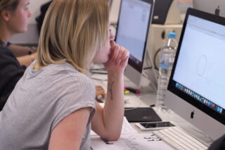 Eine Person sitzt an einem Schreibtisch, nutzt einen iMac-Computer und konzentriert sich auf ein digitales Design, das auf dem Bildschirm angezeigt wird. In der Nähe befinden sich Papiere, ein Telefon und eine Wasserflasche.