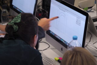 Eine Person zeigt auf einen Computerbildschirm, auf dem eine digitale Zeichnung angezeigt wird, während eine andere Person zusieht. Auf dem Schreibtisch sind Büromaterialien und Computer zu sehen.