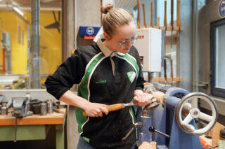 Eine Person mit Schutzbrille arbeitet an einer Drechselbank und formt mit einem Meißel ein Stück Holz in einer Werkstatt.