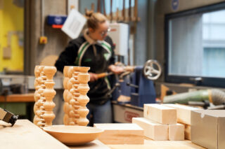 Eine Person arbeitet in einer Holzwerkstatt mit gedrechselten hölzernen Kerzenhaltern, einer hölzernen Schale und mehreren Holzblöcken auf einer Werkbank im Vordergrund.