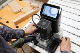 A person uses a handheld CNC routing tool with a touchscreen interface to work on a piece of wood in a workshop.