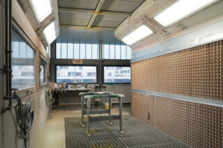 Eine saubere, leere industrielle Spritzkabine mit Metallrosten auf dem Boden, hellen Deckenleuchten und einem Arbeitstisch in der Mitte.