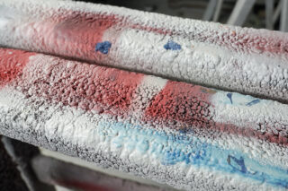 Nahaufnahme von zwei Metallrohren mit rissiger, abblätternder Farbe in den Farben Rot, Weiß und Blau und einer rauen, strukturierten Oberfläche.