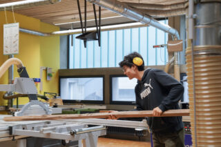 Person wearing safety earmuffs operates a woodworking machine, holding a long wooden plank in a workshop with various tools and equipment visible in the background.