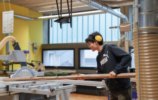 Eine Person mit Gehörschutz bedient eine Tischsäge und führt ein langes Holzbrett durch die Maschine in einer Holzwerkstatt.