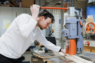 A man in a white shirt operates a large industrial saw to cut a wooden plank in a workshop.