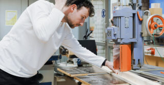 Ein Mann in einem weißen Hemd bedient eine Holzschneidemaschine und führt ein Holzbrett durch die Säge in einer Werkstatt.