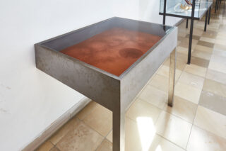 Ein Ausstellungstisch aus Metall mit Glasplatte enthält eine Schicht aus rotbraunem Pulver. Der Tisch steht auf einem gefliesten Boden neben einer weißen Wand.
