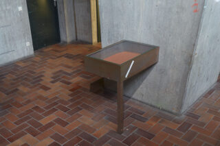 Eine Glasvitrine mit Metallrahmen steht auf einem einzelnen Bein an einer Betonwand in einem Raum mit braunem Fliesenboden. Die Vitrine besteht aus rotbraunem Material.