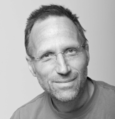 Ein Mann mittleren Alters mit kurzen Haaren, Brille und leichtem Bart, der ein legeres Hemd trägt und vor einem schlichten Hintergrund in die Kamera lächelt.