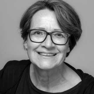 Schwarz-Weiß-Porträt einer älteren Frau mit kurzen Haaren und Brille, die in die Kamera lächelt, vor einem schlichten Hintergrund.