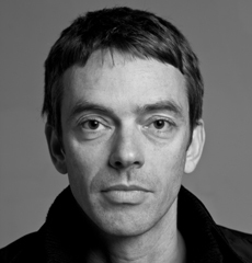 Schwarz-Weiß-Porträt eines Mannes mit kurzen Haaren, der eine dunkle Jacke trägt und mit neutralem Gesichtsausdruck direkt in die Kamera blickt.