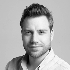 Ein Mann mit kurzen, gestylten Haaren und einem hellen Bart, der ein helles Hemd mit Kragen trägt, posiert vor einem schlichten Hintergrund.