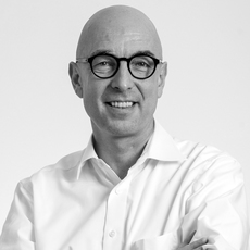 Glatzköpfiger Mann mit Brille und weißem Hemd, lächelnd mit verschränkten Armen vor einem schlichten Hintergrund.