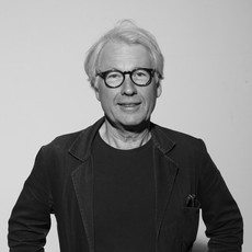 Ein Mann mit hellem Haar und Brille, der eine dunkle Jacke und ein dunkles Hemd trägt, steht vor einem schlichten Hintergrund, blickt in die Kamera und lächelt leicht.