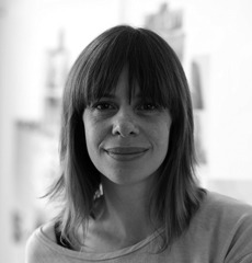 Eine Frau mit schulterlangem Haar und Pony lächelt sanft in die Kamera. Sie trägt ein helles Oberteil und steht in einem sanft beleuchteten Innenraum.