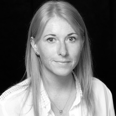 Eine Frau mit glattem, hellem Haar und einer hellen Bluse blickt vor einem dunklen Hintergrund in die Kamera.