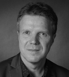 Schwarz-Weiß-Porträt eines Mannes mittleren Alters mit kurzen Haaren, der einen Blazer und ein Hemd mit Kragen trägt und direkt in die Kamera blickt.