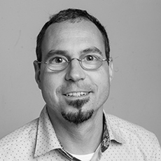 Ein Mann mit kurzen Haaren, Brille und Spitzbart lächelt in die Kamera. Er trägt ein helles, gemustertes Hemd. Der Hintergrund ist schlicht.