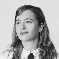 Eine Frau mit welligem Haar, Creolen und Hemd mit Kragen blickt vor einem schlichten Hintergrund leicht zur Seite.