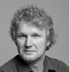 Ein Mann mit lockigem, hellem Haar und einem Hemd mit Kragen blickt vor einem schlichten Hintergrund in die Kamera.