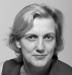 Schwarz-Weiß-Porträt einer Frau mittleren Alters mit kurzen, hellen Haaren, die ein ärmelloses Oberteil trägt und direkt in die Kamera blickt.