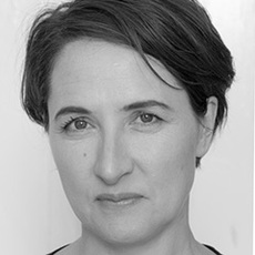 Eine Frau mit kurzen dunklen Haaren und neutralem Gesichtsausdruck, in Schwarzweiß vor einem schlichten Hintergrund fotografiert.