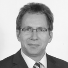 Ein Mann mit Brille, Anzug und gestreifter Krawatte blickt vor einem schlichten Hintergrund in die Kamera.