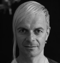 Schwarz-Weiß-Porträt einer Person mit glattem, hellem, zur Seite gekämmtem Haar, Ohrringen und direktem Blick in die Kamera vor einem dunklen Hintergrund.