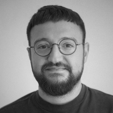 Ein Mann mit kurzen dunklen Haaren, Brille und Bart, der ein dunkles Rundhalsshirt trägt und vor einem schlichten Hintergrund in die Kamera blickt.