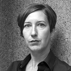 Eine Frau mit kurzem, dunklem Haar und ernstem Gesichtsausdruck steht vor einer strukturierten Steinmauer und trägt ein dunkles Hemd mit Kragen.