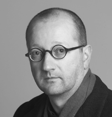 Ein Mann mit rasiertem Kopf, runder Brille und dunkler Jacke blickt vor einem schlichten Hintergrund in die Kamera.