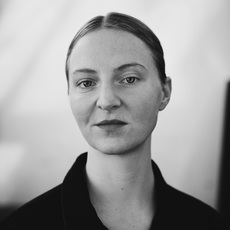 Schwarz-Weiß-Porträt einer Person mit heller Haut, kurzem, in der Mitte gescheiteltem Haar und neutralem Gesichtsausdruck, die ein dunkles Oberteil vor einem schlichten Hintergrund trägt.